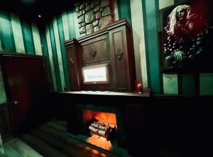 A glowing fireplace in an old-looking living room. Beside it is a large painting of an older man.