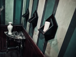 3 small black shelves, one has a white candle on it. Beyond is a small table with a vase that bears a strange symbol.