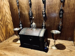 A small locked chest beside two metal cups on a shelf. Behind them are room keys hung on hooks.