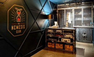 Nemesis Club lobby has their art deco logo beside a vault and an assortment of briefcases with handcuffs attached to them.