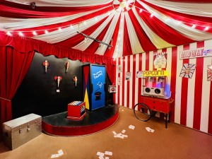 Wide angle view of the inside of a red and white circus tent. A popcorn machine sits beside a stage with props for a magic act.