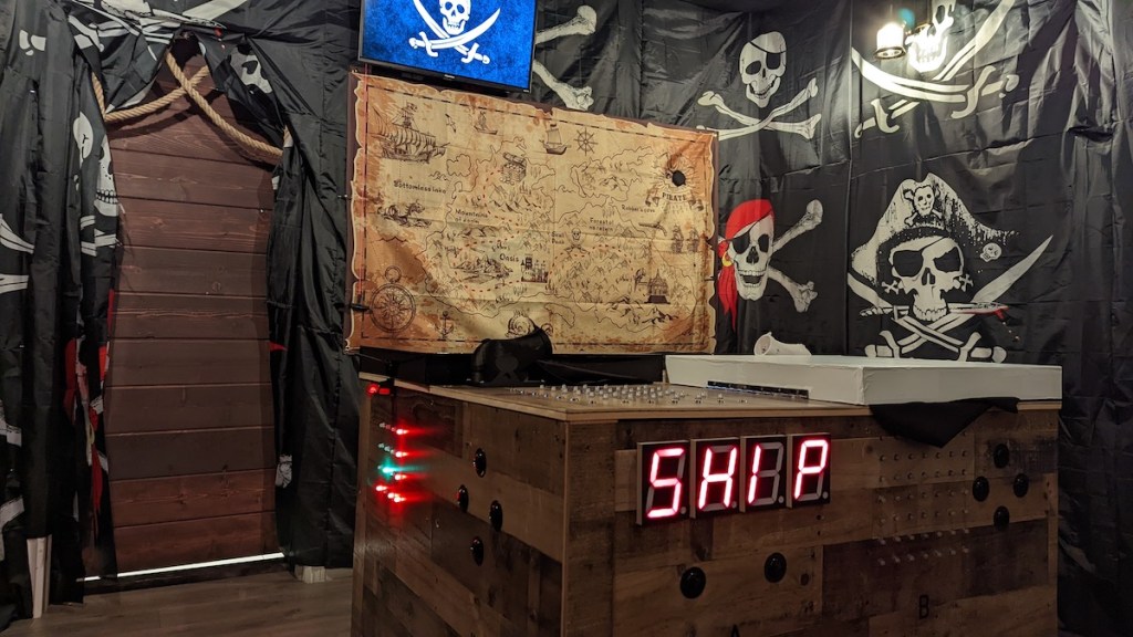 A room with walls covered in different pirate flags, in the middle is a table with a digital display that reads, "SHIP."
