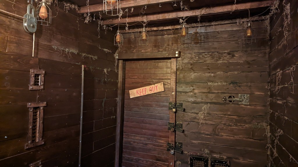Interior of an old looking wooden cabin with assorted wiring. On the far wall is a door with 3 different locks. It is labeled, "Keep Out."