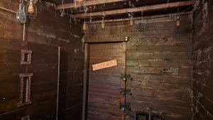 Interior of an old looking wooden cabin with assorted wiring. On the far wall is a door with 3 different locks. It is labeled, "Keep Out."