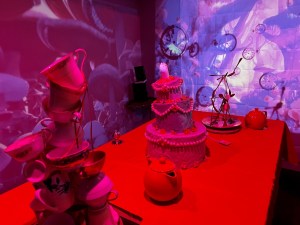 A table with a sculpture made from teacups, a tall cake, a sculpture made from spoons, and a few tea pots.