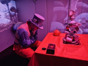 The Mad Hatter in a straightjacket sitting at the head of a table. sculpture made from teacups sits in front of him.
