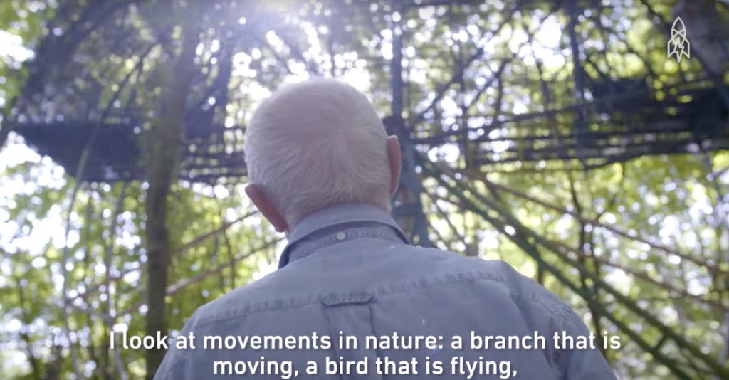 Bruno looking at a large Ferris Wheel-like structure, subtitle reads, "I look at movements in nature: a branch that is moving, a bird that is flying..."