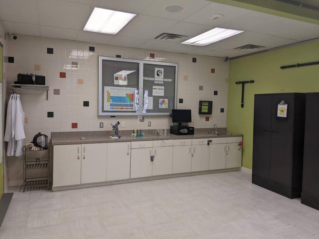 Wide angle view of a lab with man many locked cabinets, lab coats, and lab equipment.
