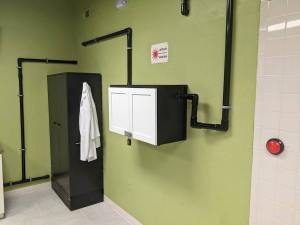 A green wall covered in black pvc tubing that connects to some larger boxes. There is a big red button and a sign warning about lasers.