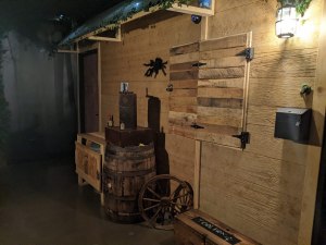 A wood cabin with an assortment of items in front including a toolbox, a wagon wheel, and a barrel.