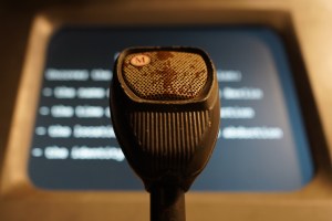 Closeup of an old microphone labeled, "M," beyond is a computer display.