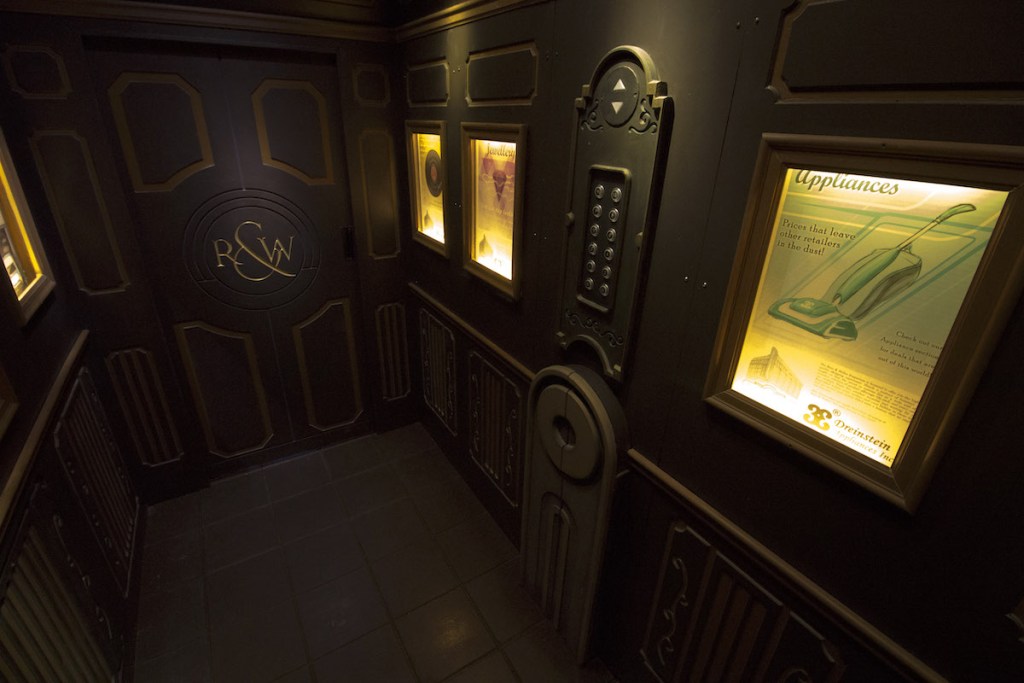 The inside of a decadent elevator with 1960s style advertisements and the letters "R&W" carved into the door.