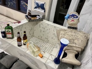 A fancy tile and marble bath that is a mess from a party, liquor and beer bottles, a bra, anda. statue of Caesar with lipstick and mask are strewn about.