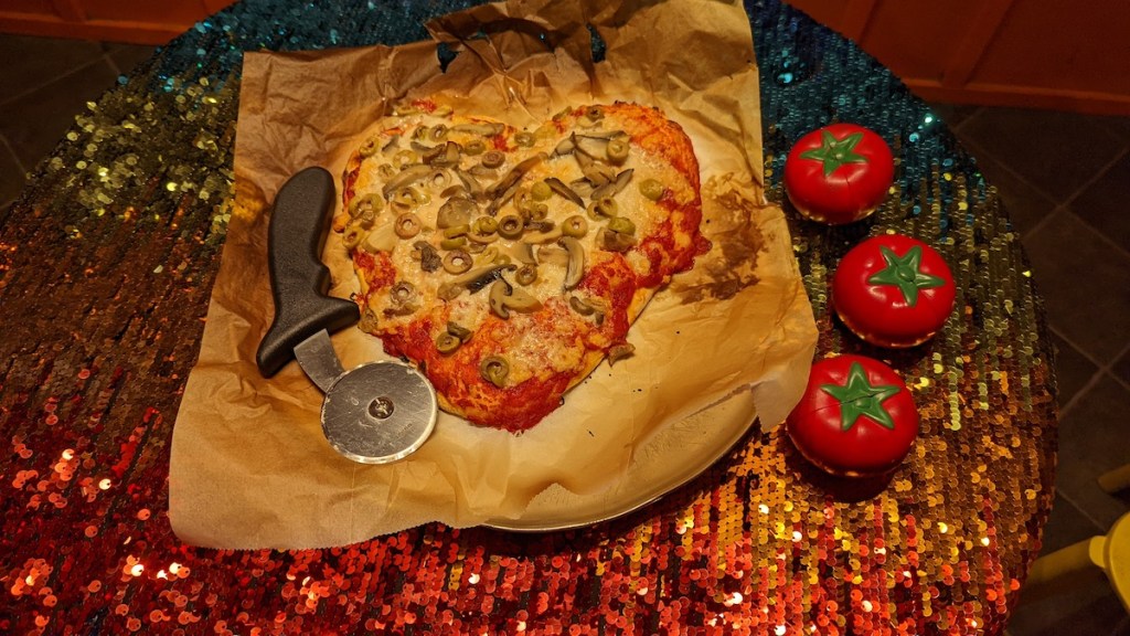 A heart-shaped pizza with a pizza cutter on one side and three tomatoes on the other.