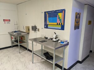 A science lab with steel tables, scientific equipment, a periodic table, and a Fallout Shelter sign on the wall.
