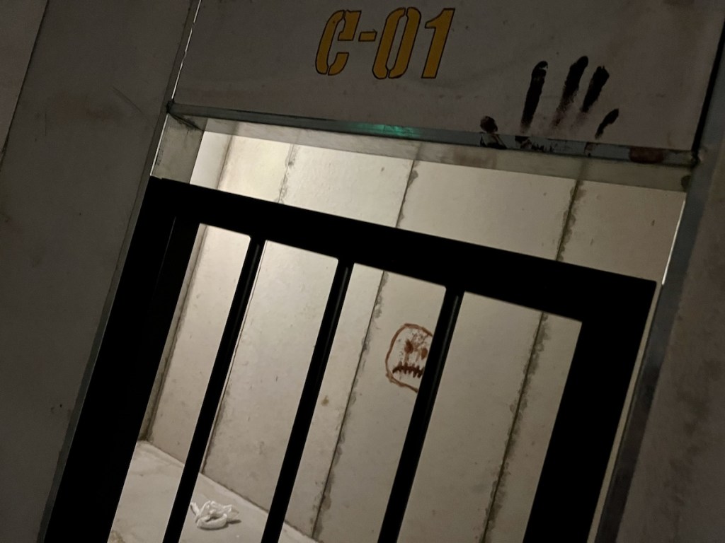 A prison cell labeled "C-01" there is a bloody handprint on the wall above it, and a small piece of toilet paper on a bench inside of the cell.