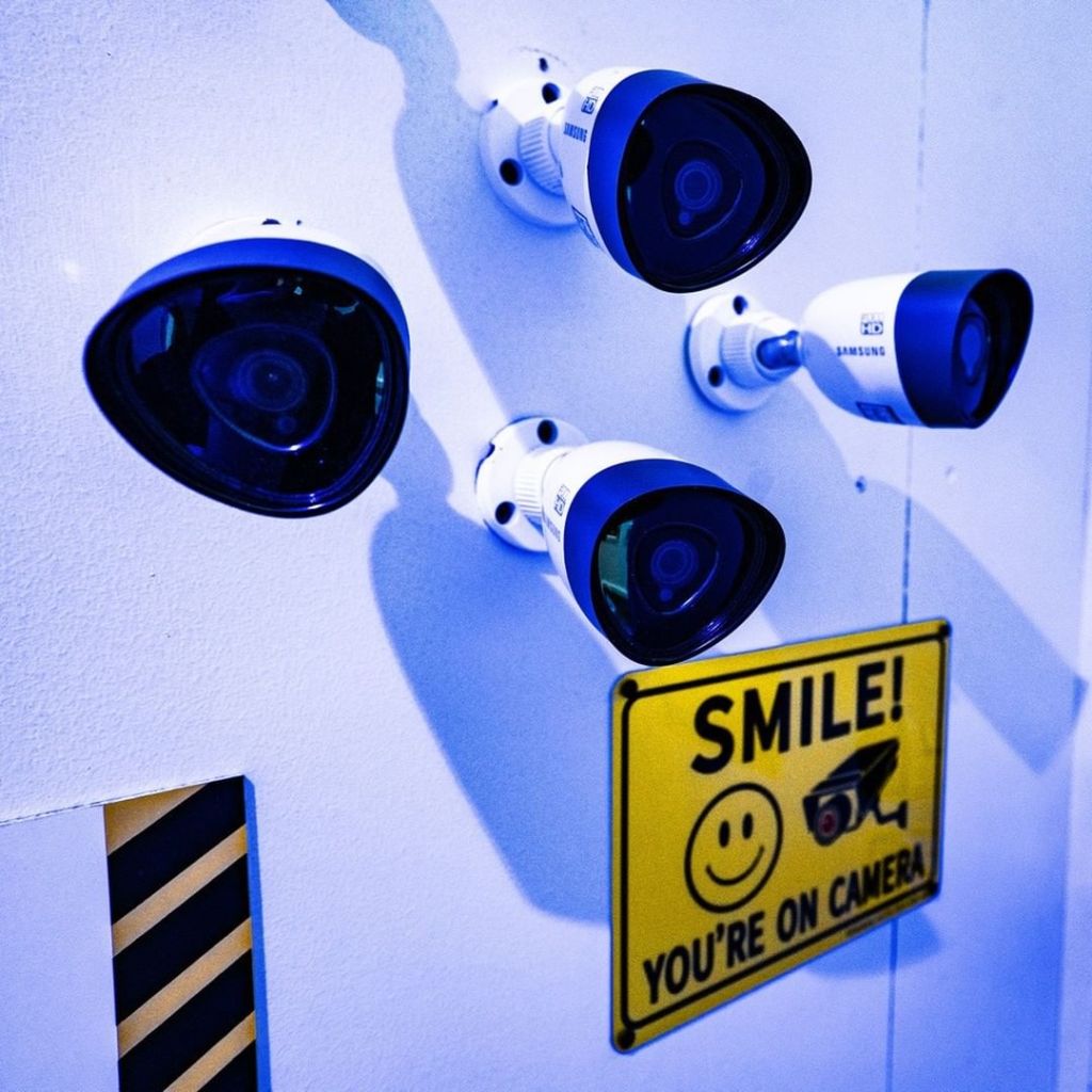A wall of security cameras with a yellow sign that reads, "SMILE! You're on camera"