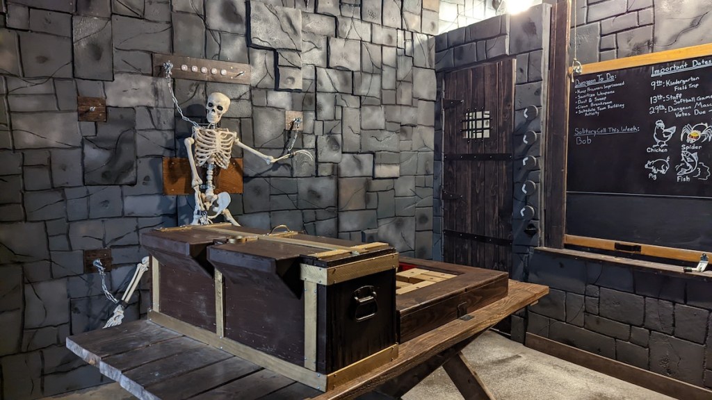 A dungeon with a skeleton hung from the wall, a large wooden contraption, and a blackboard with writing.