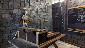 A dungeon with a skeleton hung from the wall, a large wooden contraption, and a blackboard with writing.