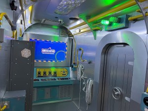 Wide angle view of a submarine, it's long and narrow, and featuring a control panel, lockers, and a big metal door.