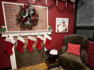 A large chair beside a fireplace decorated for Christmas with stockings, a wreath, elves, and a picture of Santa.