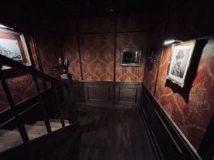 The hallway and staircase in an old hotel. On one wall is a portrait of HH Holmes, opposite is a photo of the White City - the 1893 World's Fair in Chicago.