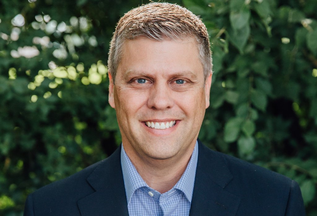man with short neat grey/brown hair, blue eyes, smiling, in a checkered shirt, blue blazer, against a background of greenery