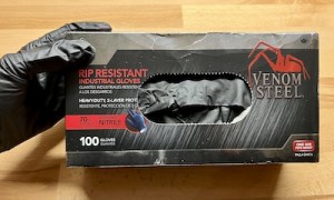 A hand wearing a black glove holding a box of 100 Venom Steel gloves.