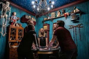 A seance parlor with 5 people standing around a table looking upwards with wonder in their eyes.