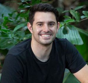 caucasian man smiling, with neat brown hair, wearing a black t-shirt, in front of greenery.