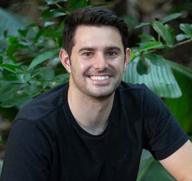 caucasian man smiling, with neat brown hair, wearing a black t-shirt, in front of greenery.