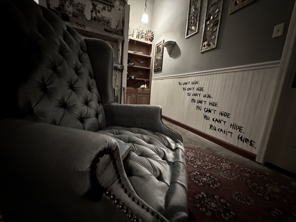 A large chair in a living room, beyond it is a wall that has the words, "You can't hide" written repeatedly on it.