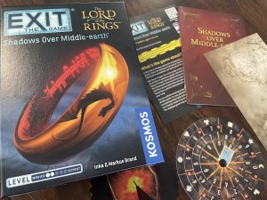 EXIT's Lord of the Rings Shadows Over Middle-earth game box with the One Ring. Beside it are assorted game items including instructions, story pamphlet, a map, the Eye of Sauron, and a decoder wheel.