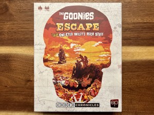 The Goonies: Escape with One-Eyed Willy’s Rich Stuff's box depicts an orange skull, within the skill is an illustration of the Goonies, a pirate ship, and a large amount of treasure.