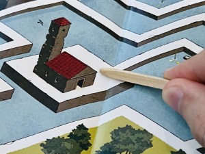 Closeup of a maze, fingers hold a small pointed stick to keep track of our place within the maze.