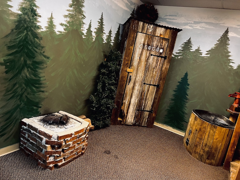 An outhouse with odd symbols between a brick fire pit, and a wooden well.