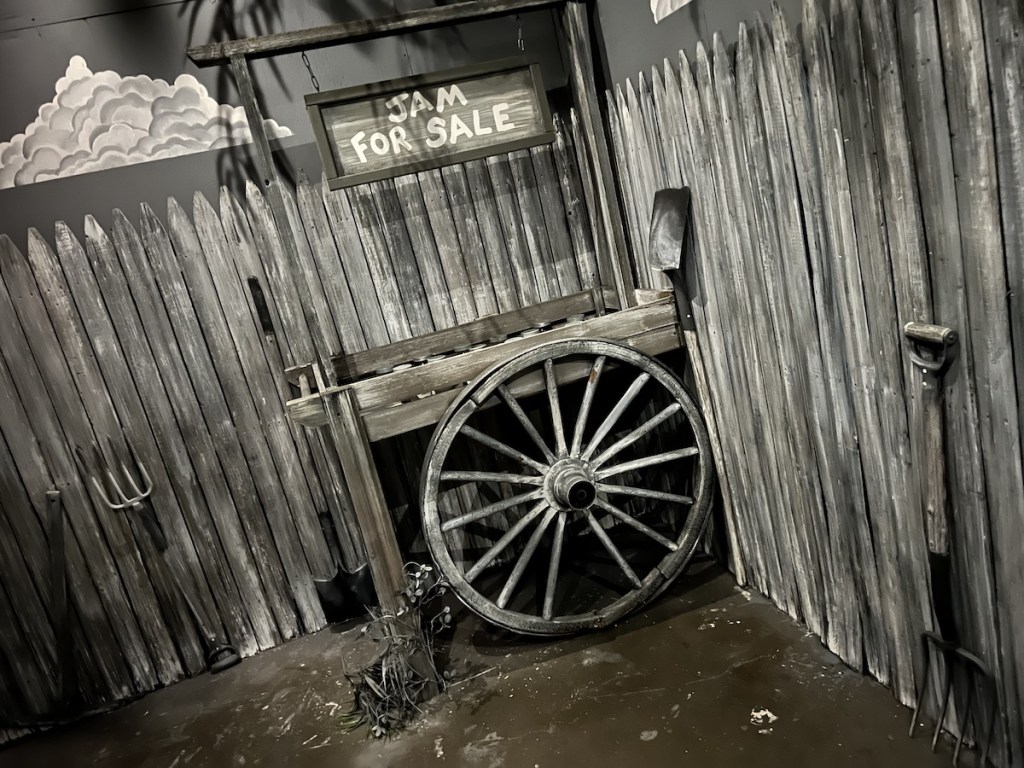 A black & white set featuring a cart selling jam, surrounded by a fence and farming tools.