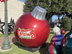 A gigantic Christmas ornament on lawn in a park surrounded by smaller oversized ornaments. It is labeled, "The Grapevine Texas Christmas Capital of Texas."