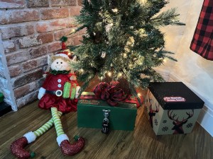 A plush elf holding a package under a small Christmas tree beside a locked box.