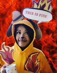 asian woman dressed up as the "this is fine" meme, in orange hoodie with cartoon dog ears and hat, paper caption that says "this is fine" and paper flames, holding a mug against a backdrop of flames