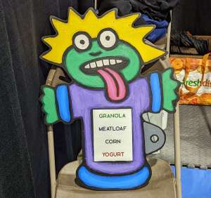 Alien cardboard standee whose shirt reads, "GRANOLA MEATLOAF CORN YOGURT"