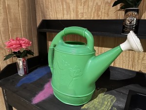 A large green watering can on a workbench with flowers planted in empty beer and soda cans.