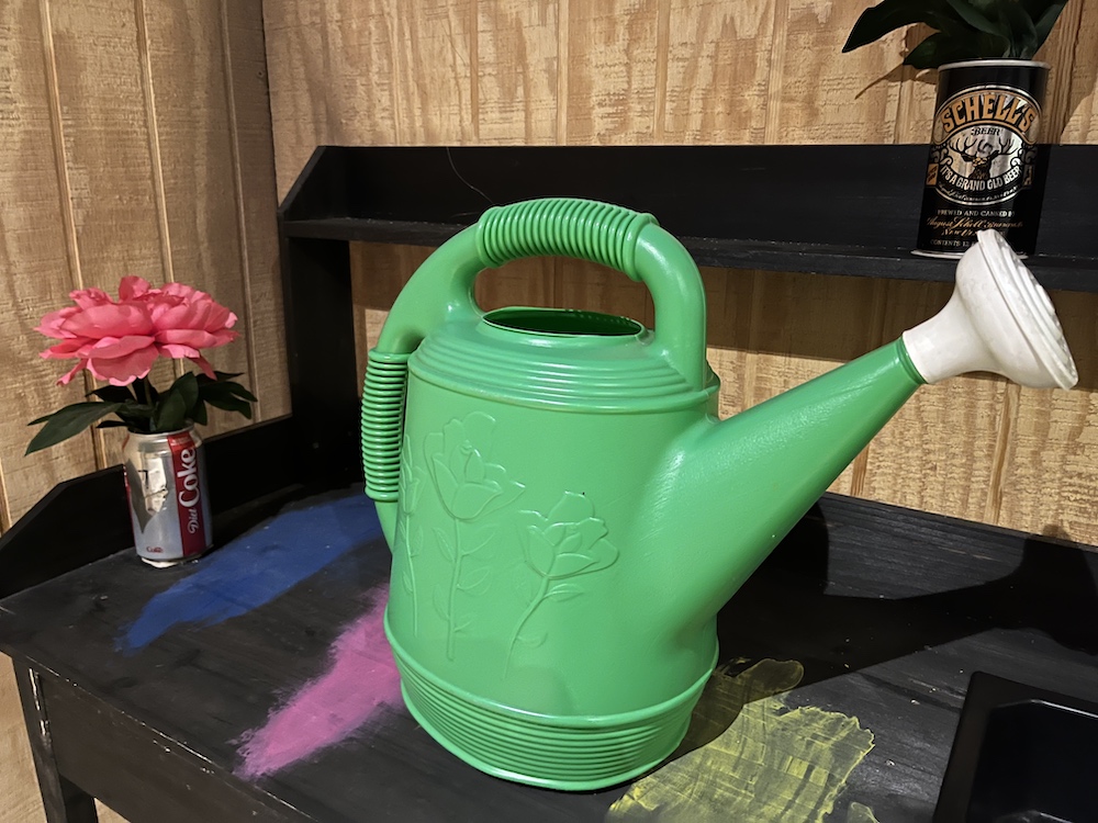 A large green watering can on a workbench with flowers planted in empty beer and soda cans.
