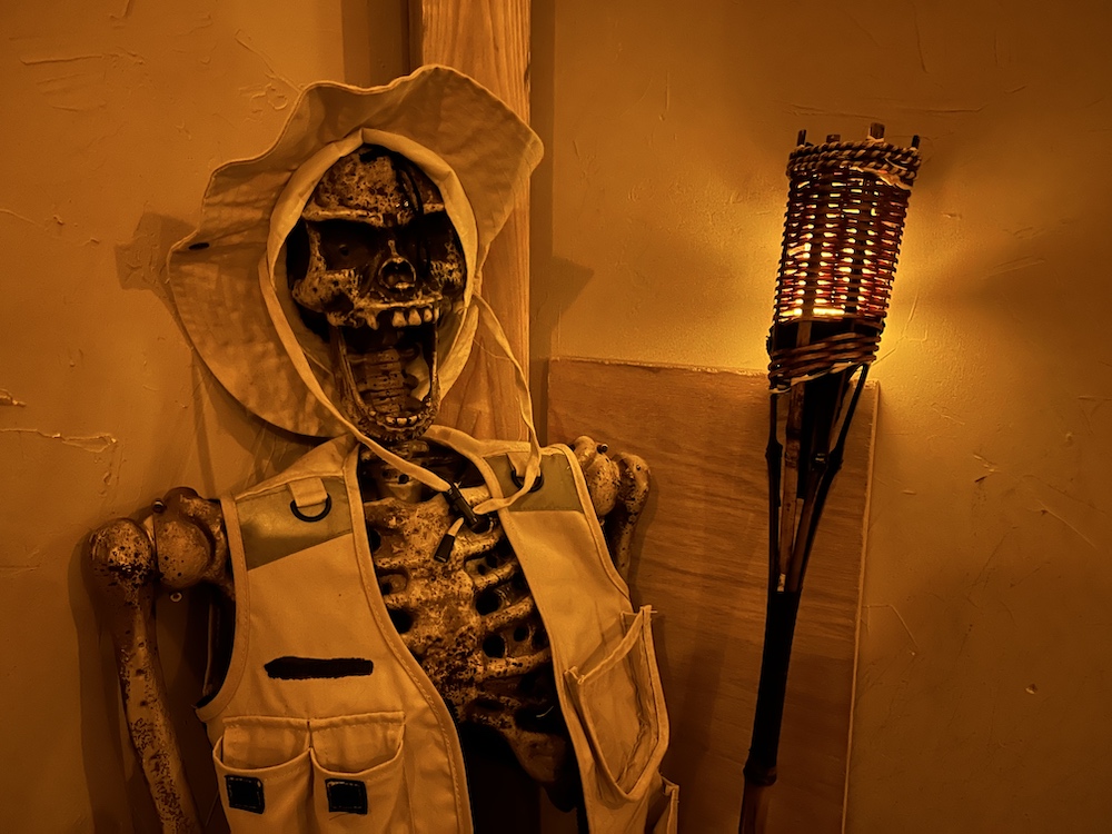 A halloween prop skeleton in a tan vest and hat holding a tiki touch.
