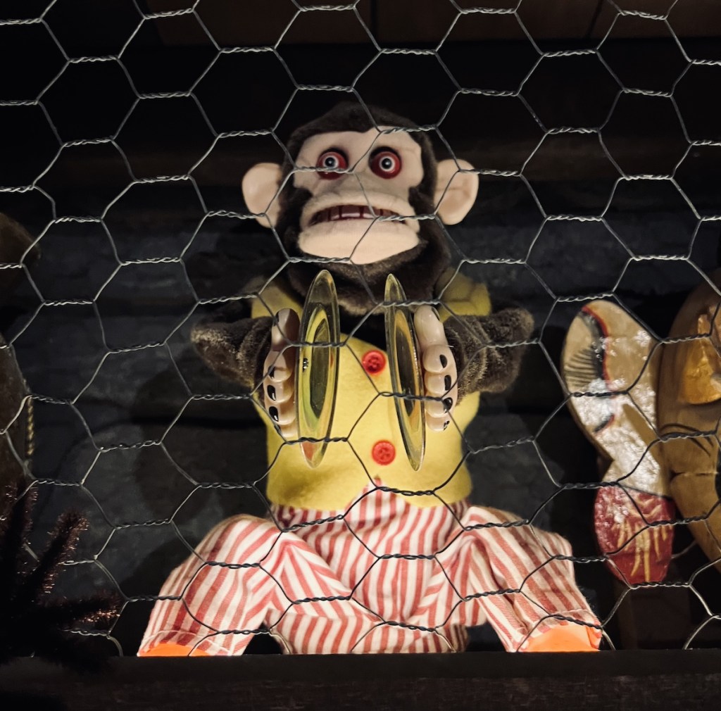 A creepy monkey toy holding symbols behind chickenwire, staring into the camera.