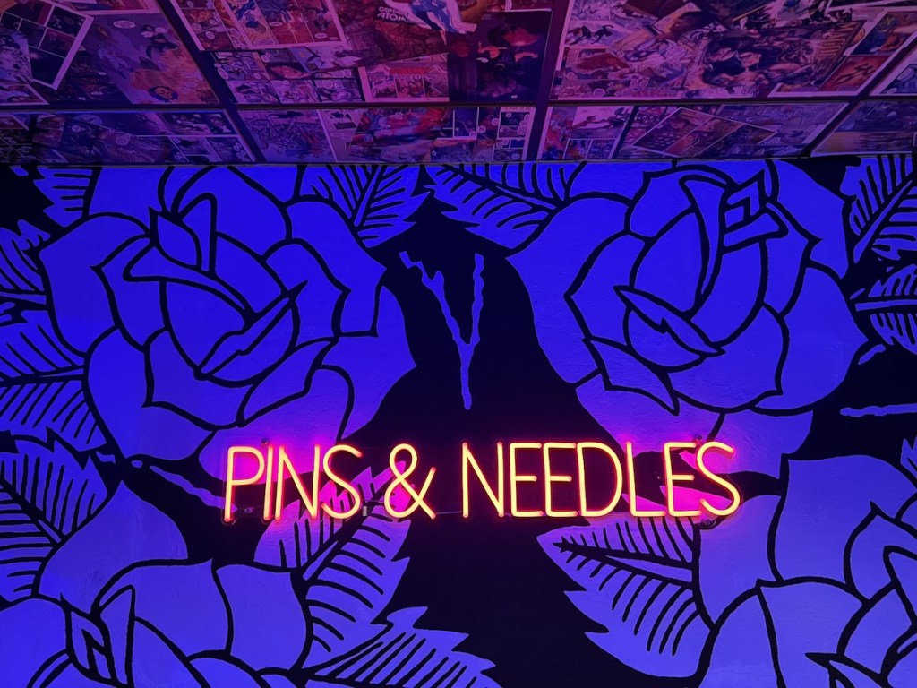 The words "PINS & NEEDLES" glowing in red neon against a wall papered in black & white roses.