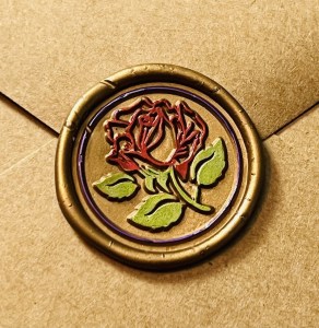 Closeup of a rose wax seal that is touched up with colored markers adding red to the rose, green to the leaves, and a purple ring around it all.