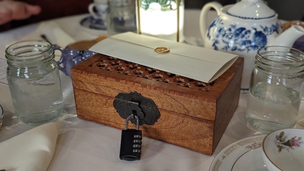 A beautiful wooden box with a padlock. Atop it is a letter wax sealed in gold. It is surrounded by tea settings.