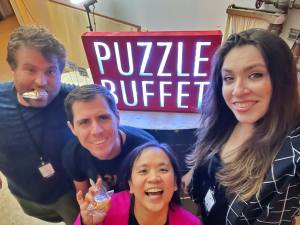 Cara, Tammy, Brendan, and Ryan holding up fortune cookies in front of an illuminated "Puzzle Buffet" sign.