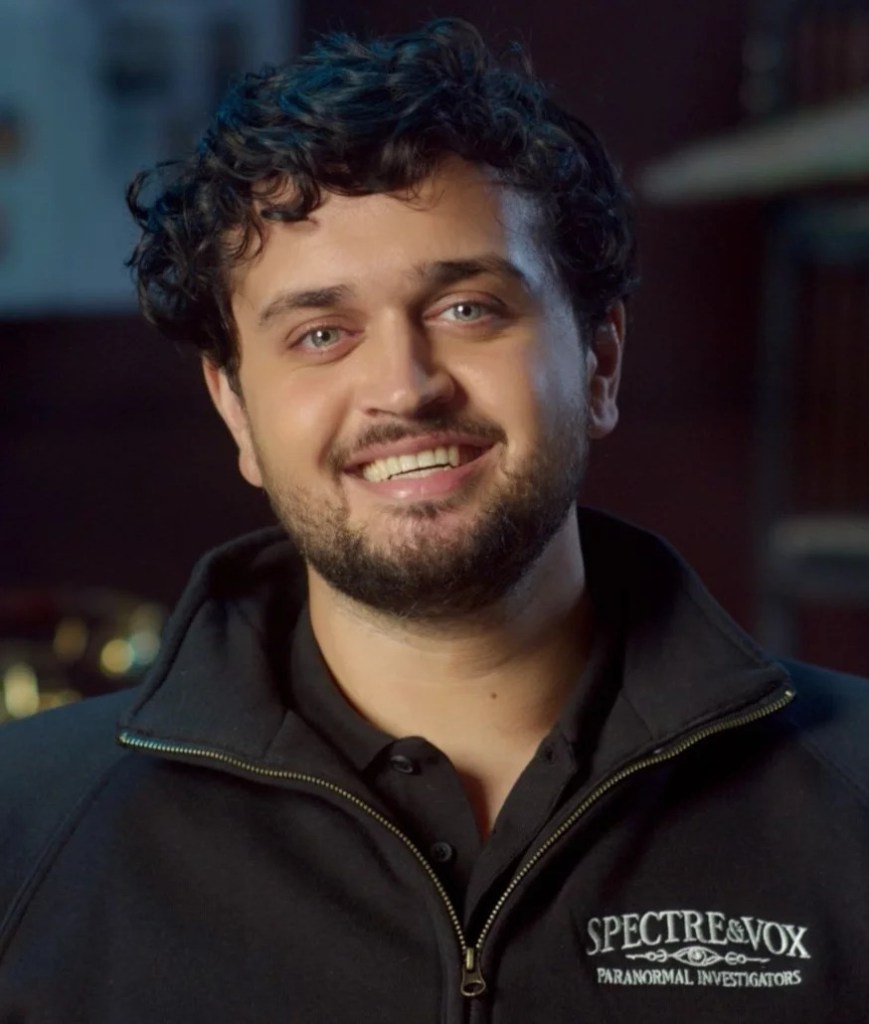 caucasian man with curly brown hair, short beard, and light blue eyes, smiling rakishly while wearing a black zip up sweatshirt that says "spectre & vox, paranormal investigators""